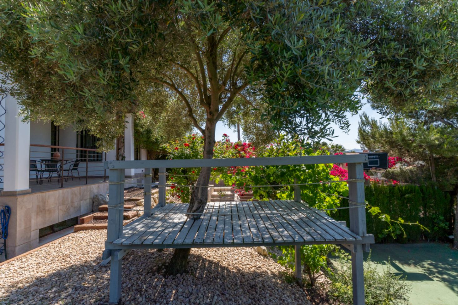 Plataforma de madera integrada alrededor de un olivo en el jardín, con vistas a la terraza principal y zona de juegos con canasta.
