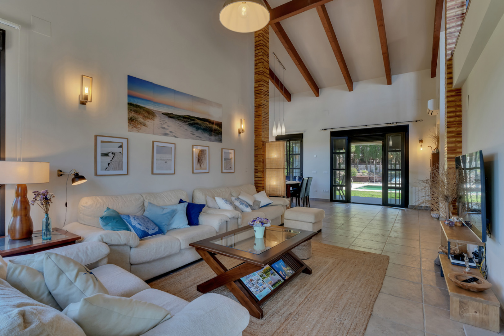 Salón comedor con techos de doble altura y vigas de madera, suelos de gres y grandes puertas acristaladas que conectan con el jardín y la piscina.