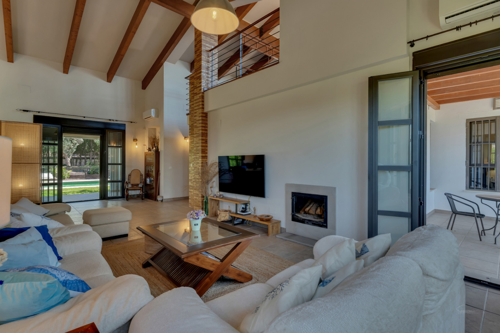 Salón de techos altos con vigas de madera, chimenea integrada y acceso directo al jardín y terraza a través de amplios ventanales.