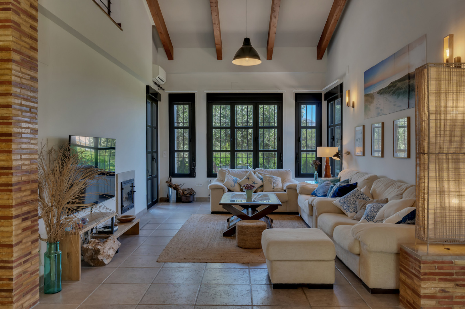 Salón elegante con techos altos y vigas de madera expuestas. Cuenta con chimenea integrada, suelos cerámicos y amplios ventanales con marcos negros que aportan luz natural.