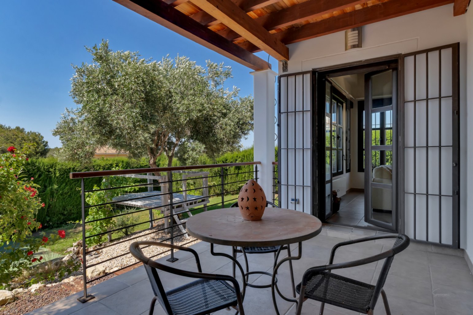 Terraza cubierta con vigas de madera vista, zona de estar exterior y vistas despejadas al jardín con olivo y setos de privacidad.