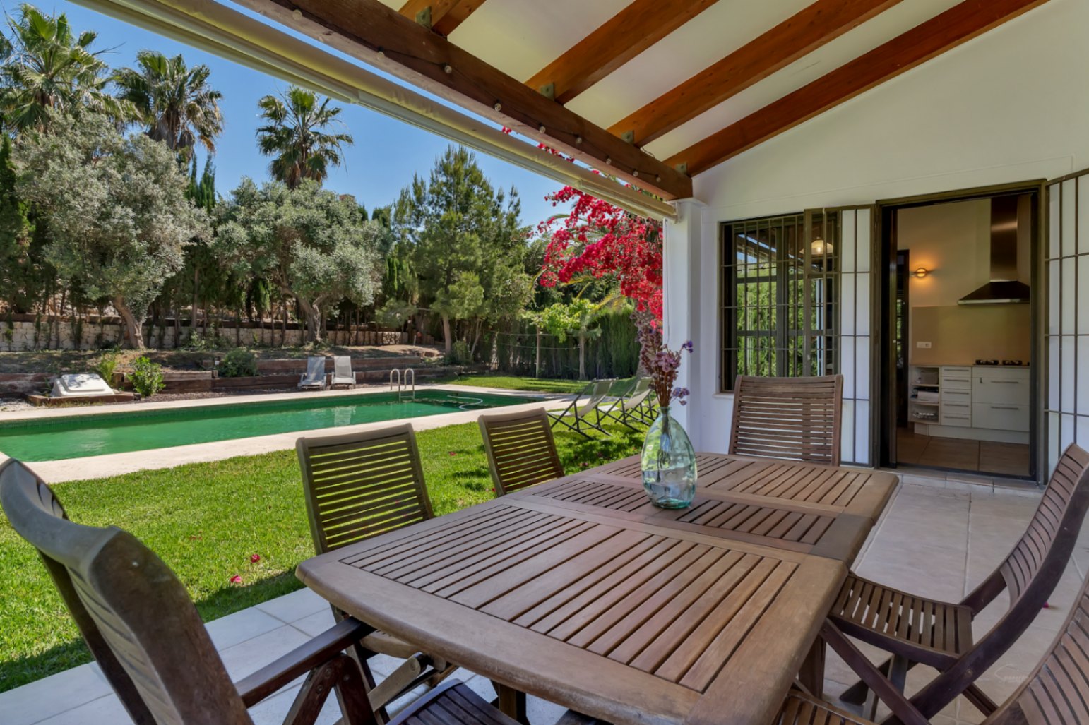 Terraza cubierta con vigas de madera y zona de comedor exterior, con vistas directas al jardín, la piscina y acceso inmediato a la cocina de la vivienda.
