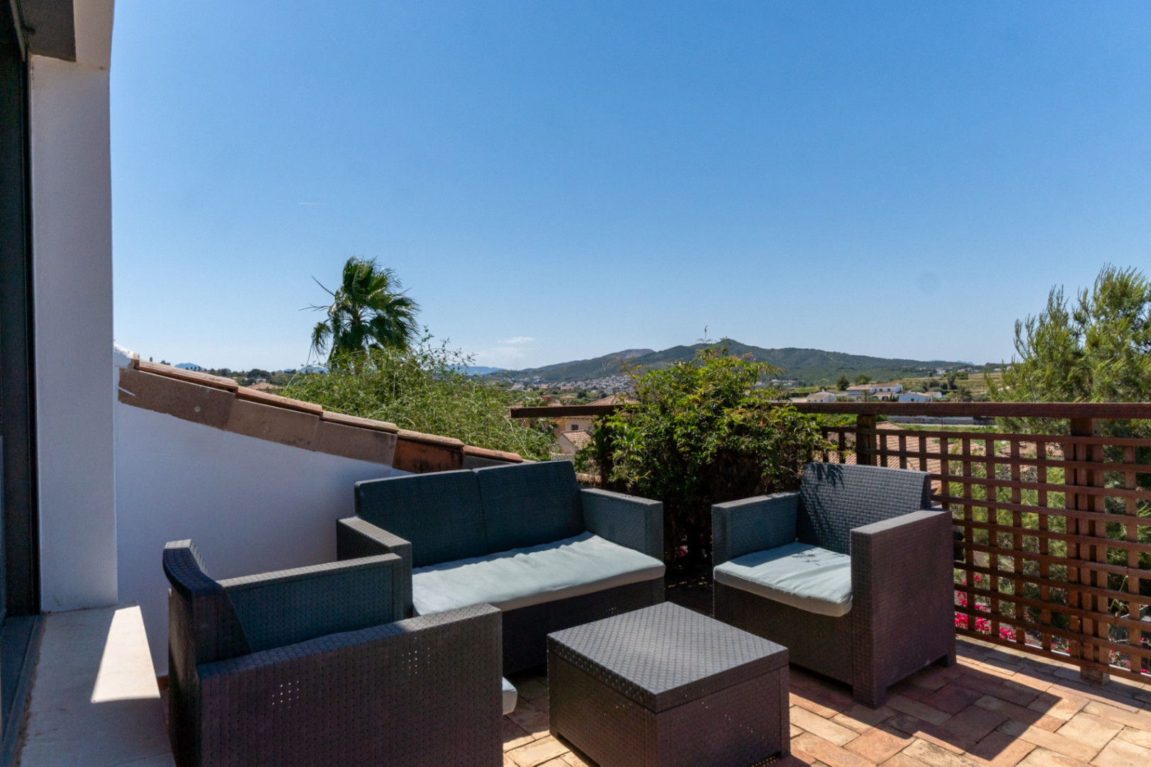 Terraza superior con mobiliario de ratán sintético, suelo de barro cocido y vistas despejadas a las montañas y el entorno de Jávea bajo un cielo despejado.