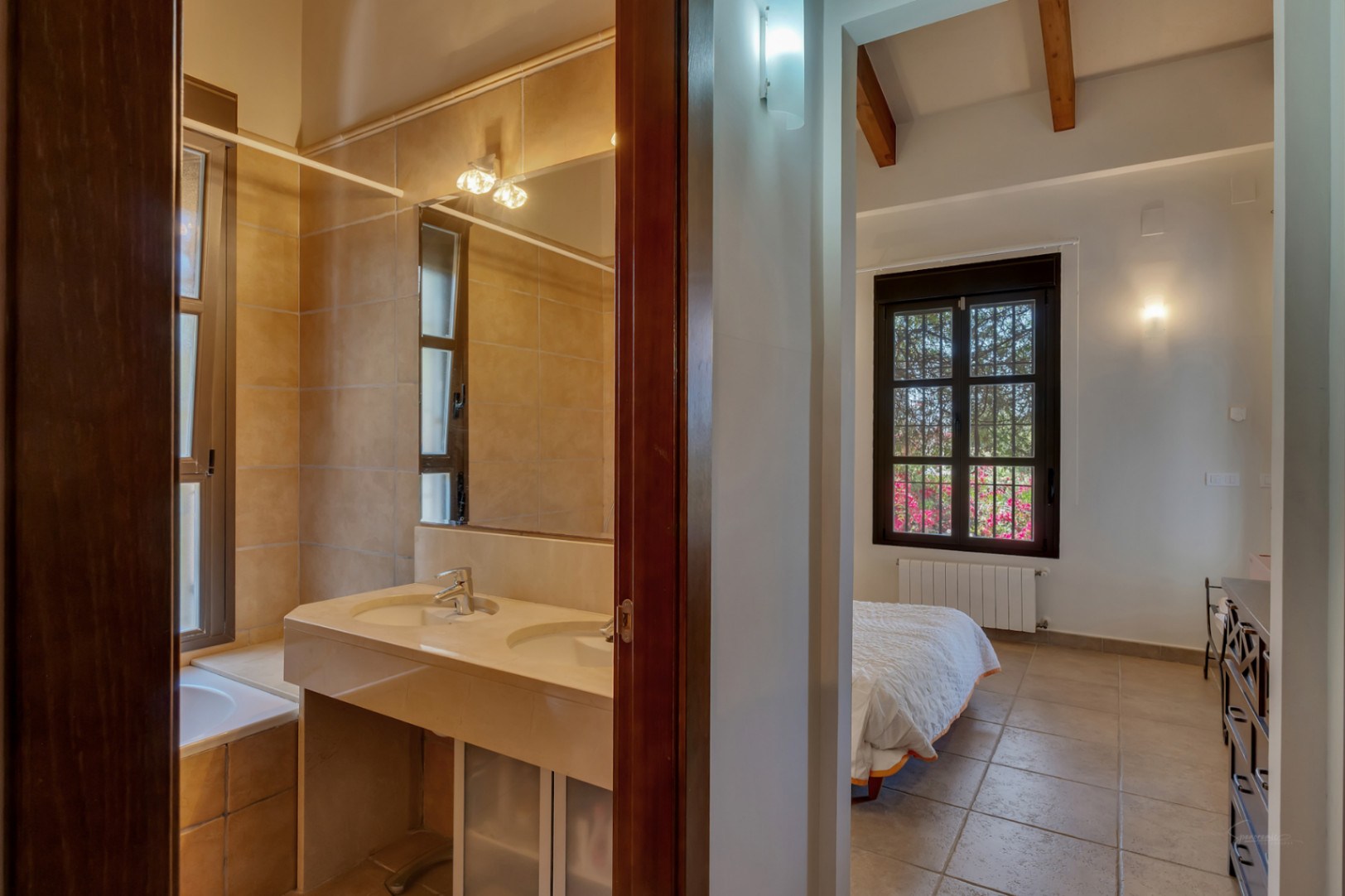 Vista conectada entre un baño con lavabo doble de piedra y un dormitorio con vigas de madera, suelos cerámicos y ventana al jardín.