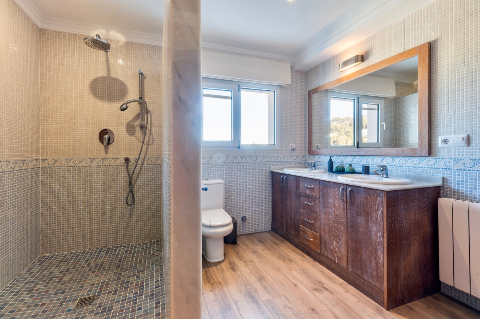 Baño con ducha de mosaico, mueble de madera de doble seno y suelos de efecto madera. La estancia cuenta con luz natural y acabados en azulejo tradicional.