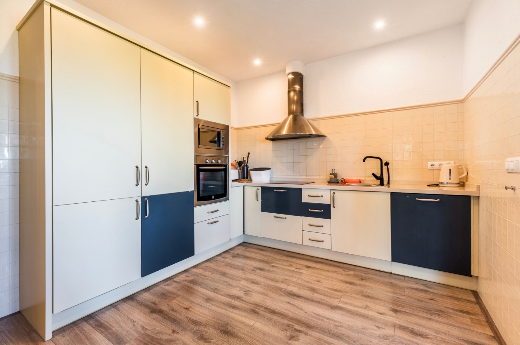Cocina moderna con armarios en tonos crema y azul, equipada con horno y microondas encastrados, campana de acero y suelo de efecto madera.