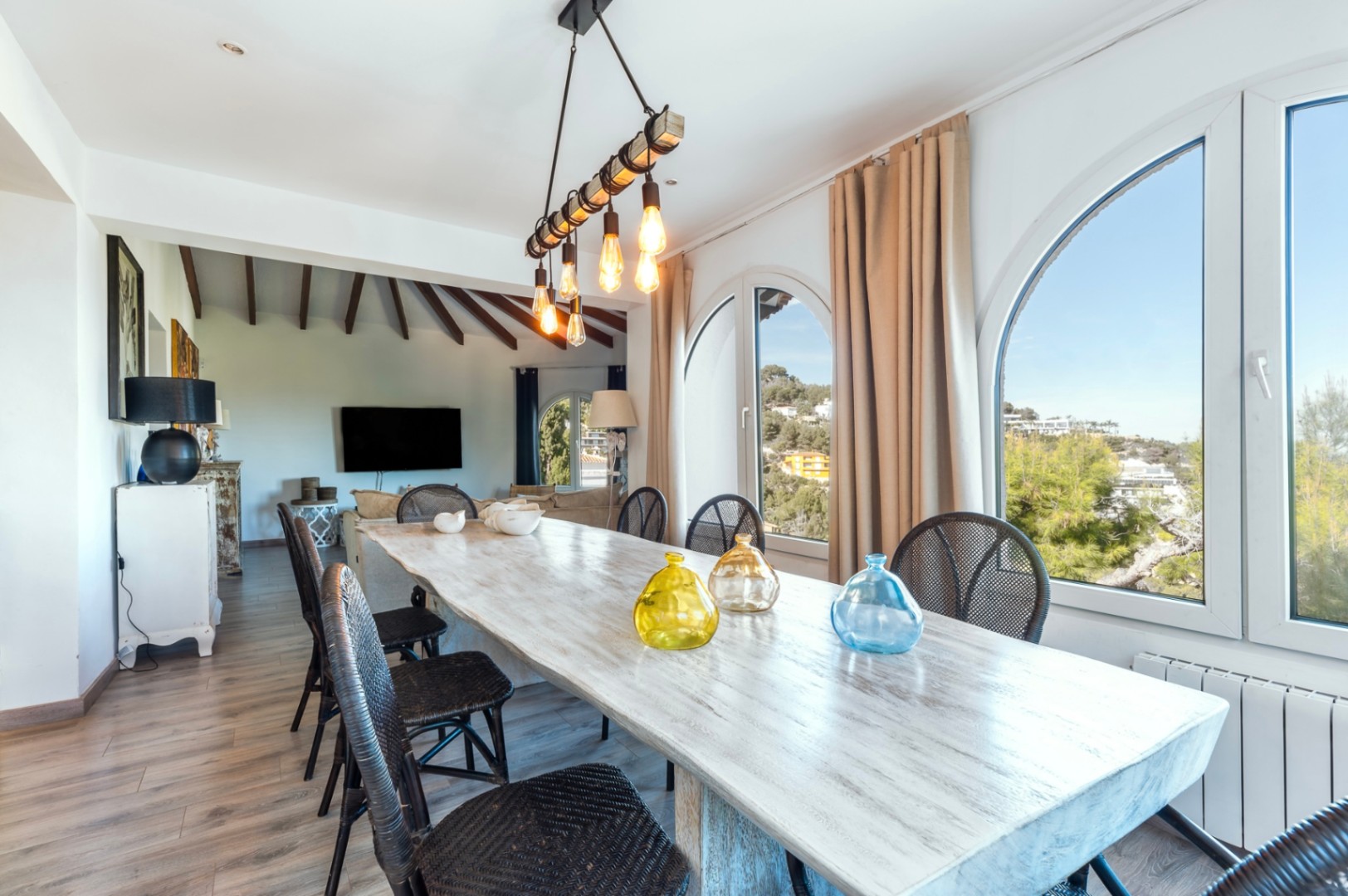 Comedor luminoso con mesa de madera decapada, sillas de mimbre y grandes ventanales en arco con vistas despejadas al entorno natural de Jávea.