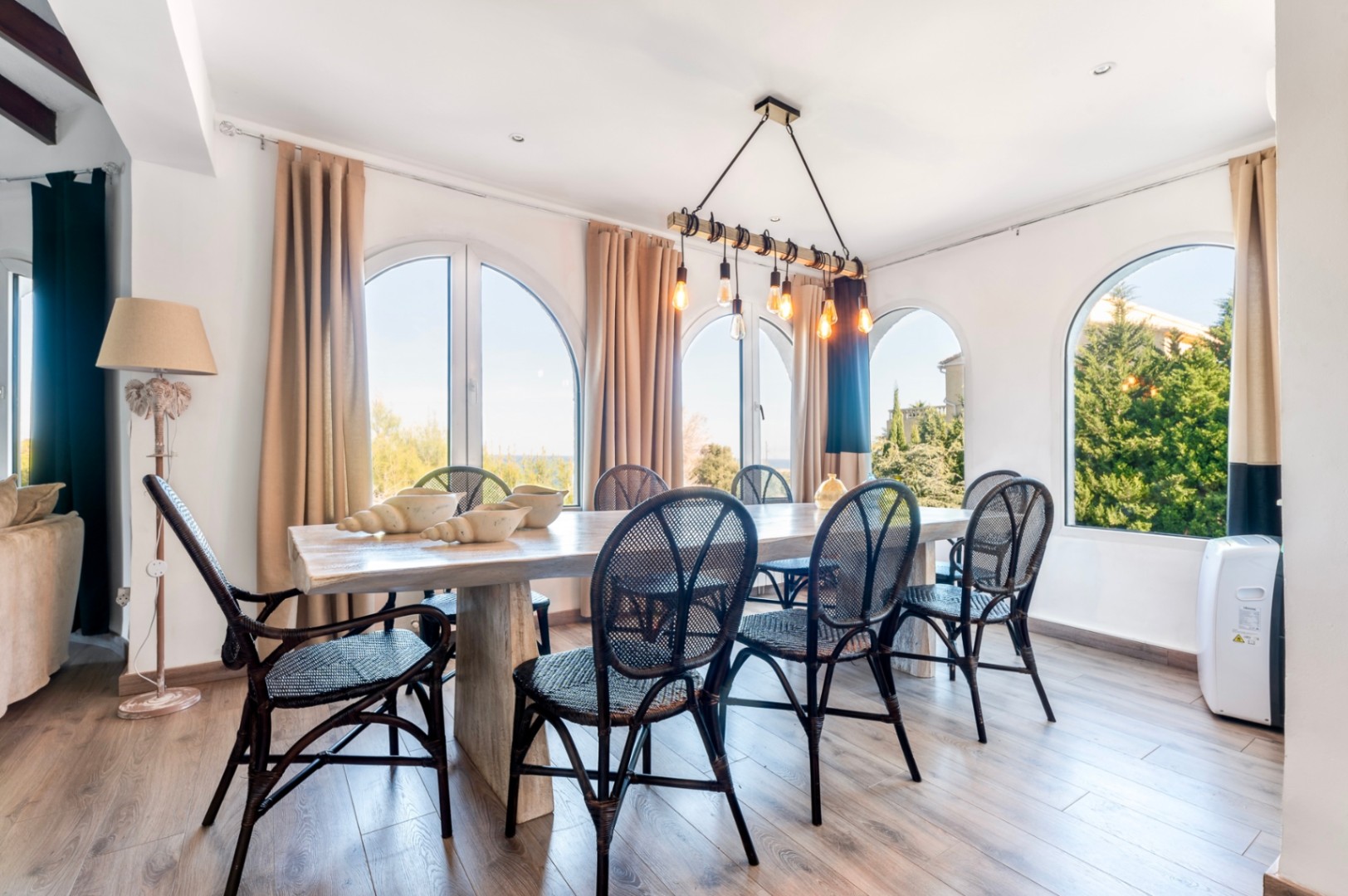 Comedor luminoso con ventanales de arco, mesa de madera maciza y sillas de mimbre negro bajo una lámpara colgante de estilo industrial.