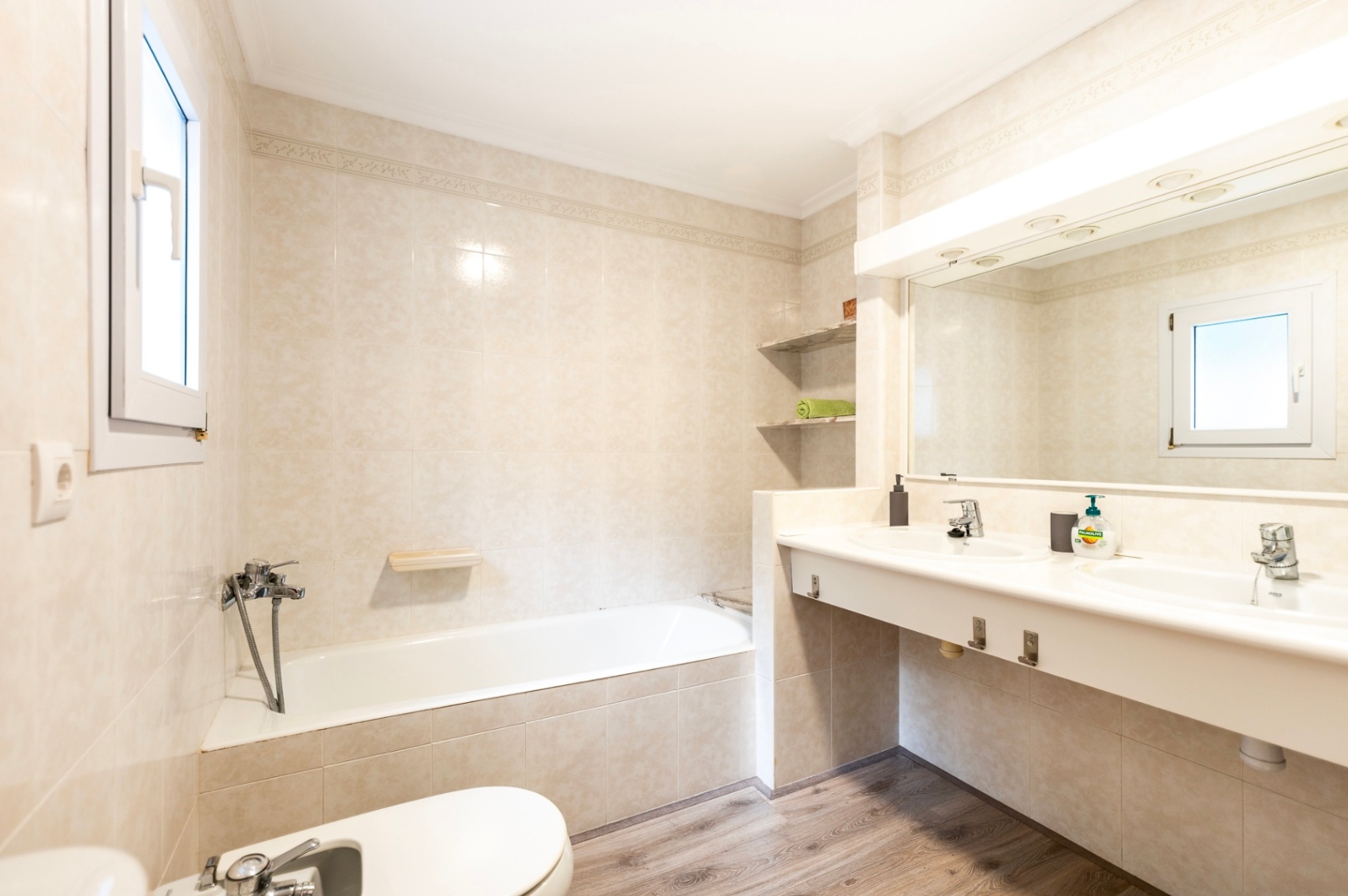 Cuarto de baño con bañera integrada, lavabo doble encastrado y suelos de efecto madera. Incluye azulejos en tonos crema, bidet y un amplio espejo con iluminación superior.