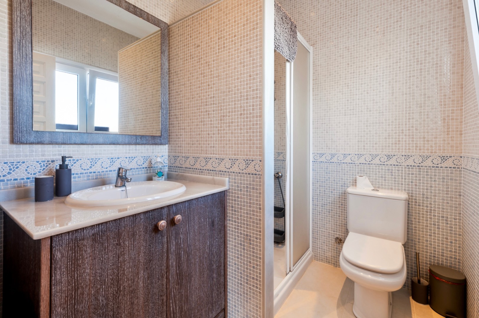 Cuarto de baño con paredes de mosaico en tonos beige y cenefa azul. Incluye un mueble de lavabo en madera oscura con encimera de mármol y ducha con mampara.