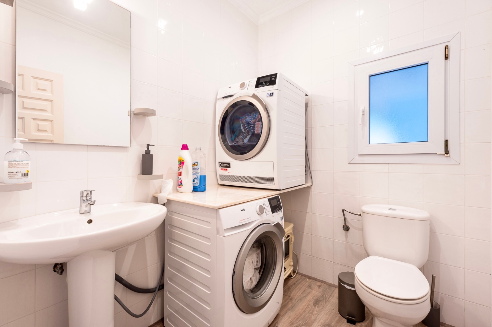 Cuarto de lavado y aseo funcional con paredes alicatadas en blanco, equipado con lavadora y secadora AEG, lavabo de pedestal y suelos de efecto madera.