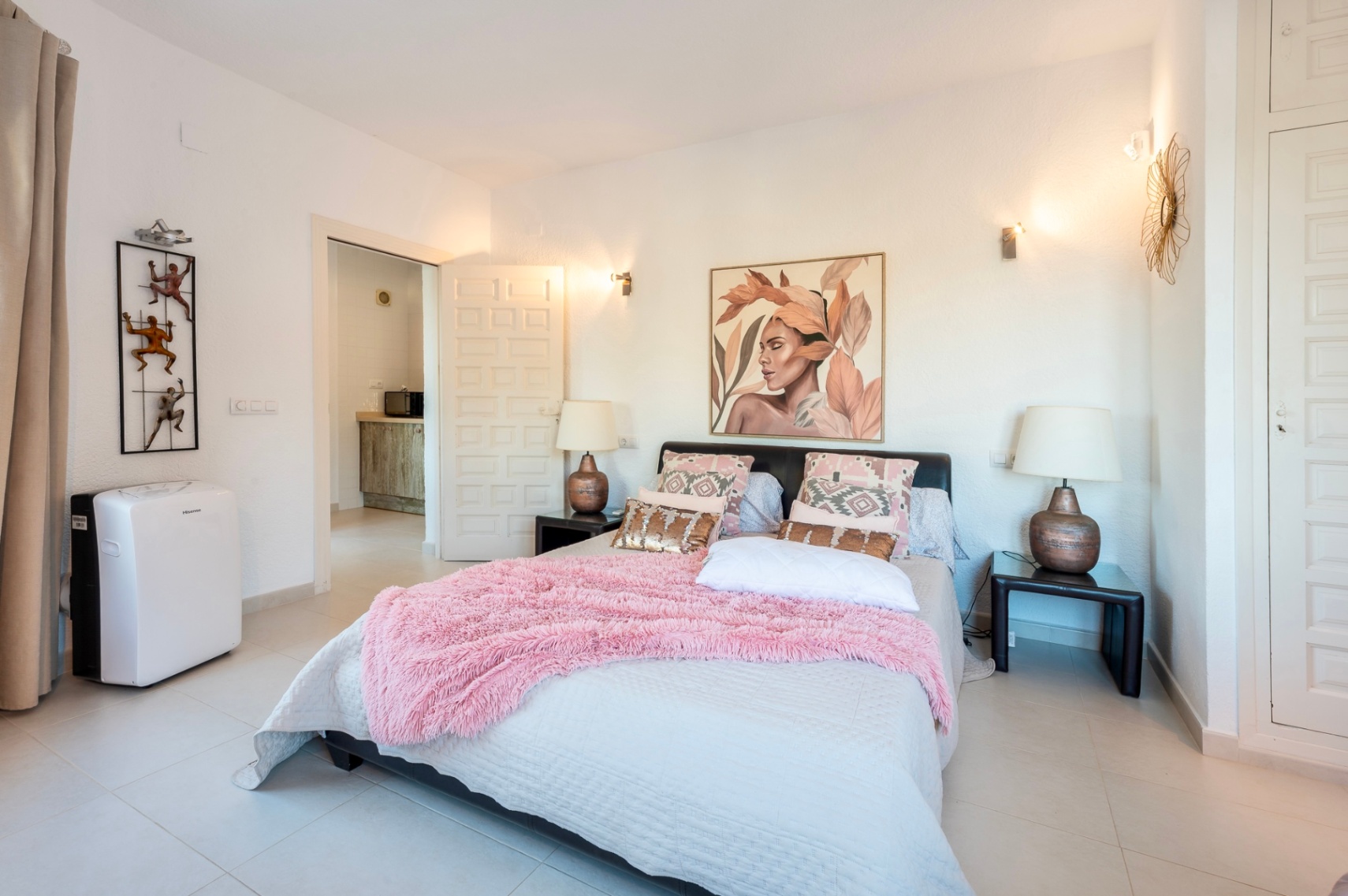 Dormitorio con cama doble, textiles rosas y armarios empotrados. Presenta suelos cerámicos claros, iluminación mediante apliques y una unidad de aire acondicionado portátil.