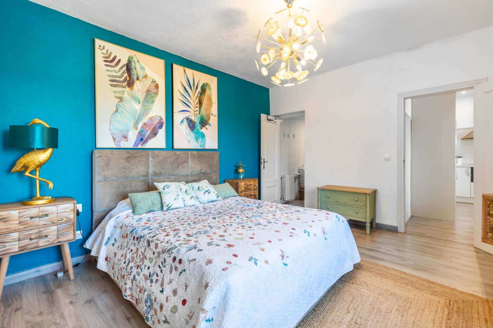 Dormitorio con pared de acento turquesa, cabecero tapizado en gris y cuadros botánicos. Destaca la lámpara dorada de diseño, el suelo de madera clara y la iluminación moderna.