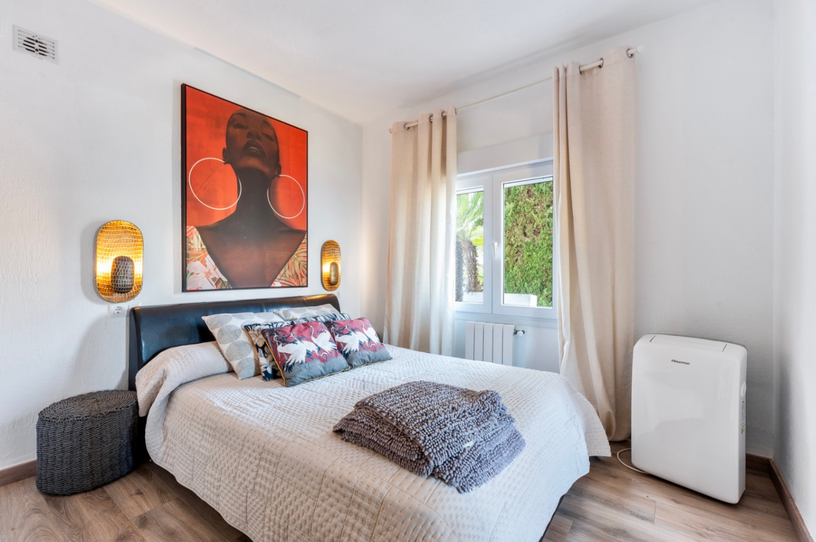 Dormitorio luminoso con suelos de madera, cabecero de cuero oscuro y un gran cuadro decorativo. Incluye apliques dorados, radiador y una unidad de aire acondicionado portátil.