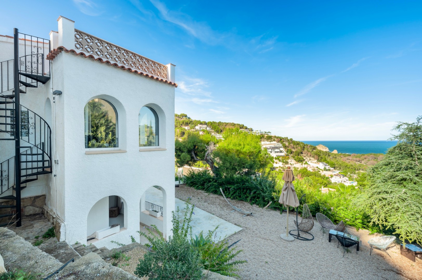 Fachada blanca con arcos tradicionales, escalera de caracol metálica y zona de estar exterior sobre grava con vistas panorámicas al mar Mediterráneo.