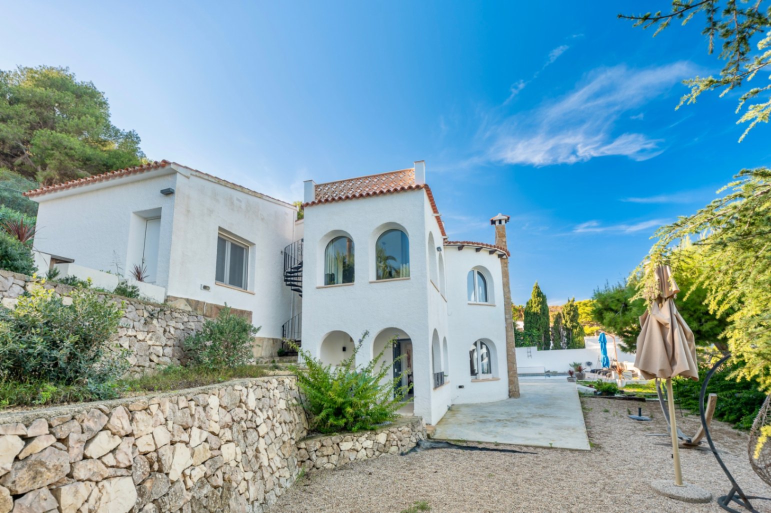 Fachada blanca de estilo mediterráneo con arcos tradicionales, muros de piedra natural y escalera de caracol exterior bajo un cielo despejado en Jávea.