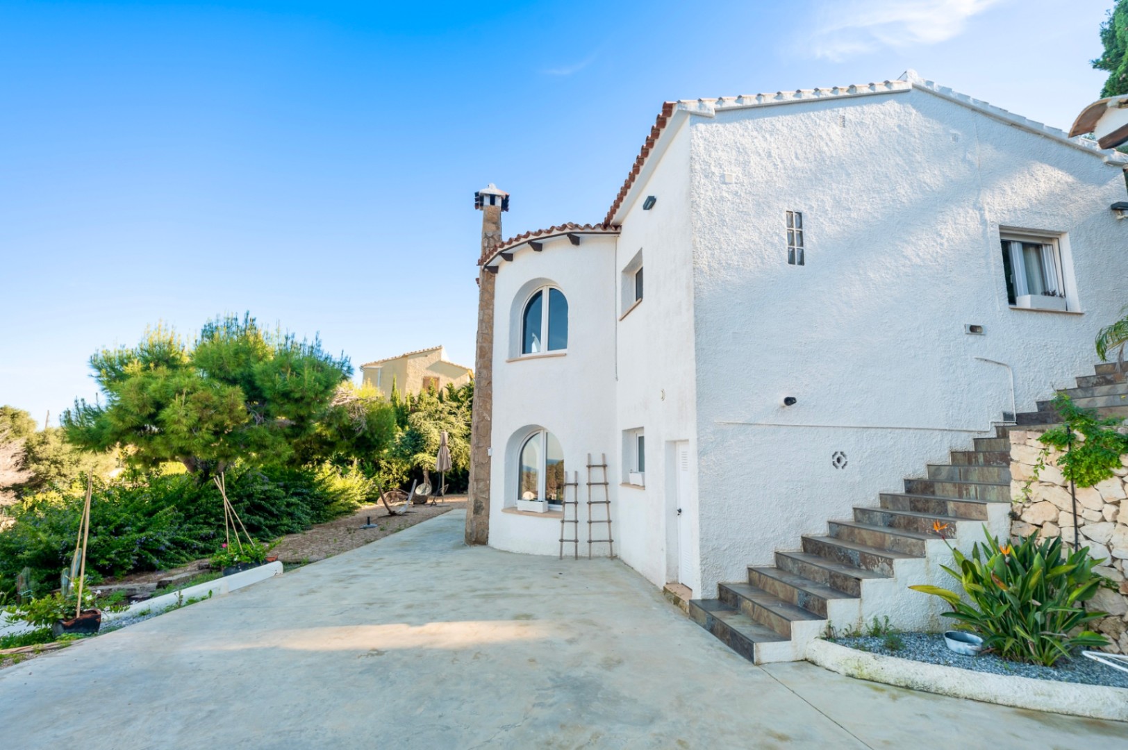 Fachada mediterránea con muros blancos, ventanas de arco y chimenea de piedra, junto a una escalera exterior que conecta los niveles de la propiedad.