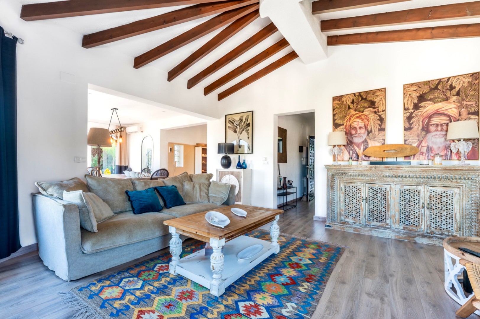 Salón luminoso con techos de vigas de madera vista, mobiliario de estilo rústico y un aparador tallado. La estancia integra una zona de estar con suelos de madera y decoración étnica.