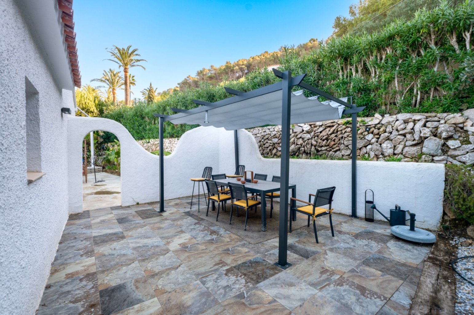 Terraza exterior con comedor bajo pérgola moderna, suelos de piedra natural y muros blancos tradicionales junto a una ladera ajardinada.