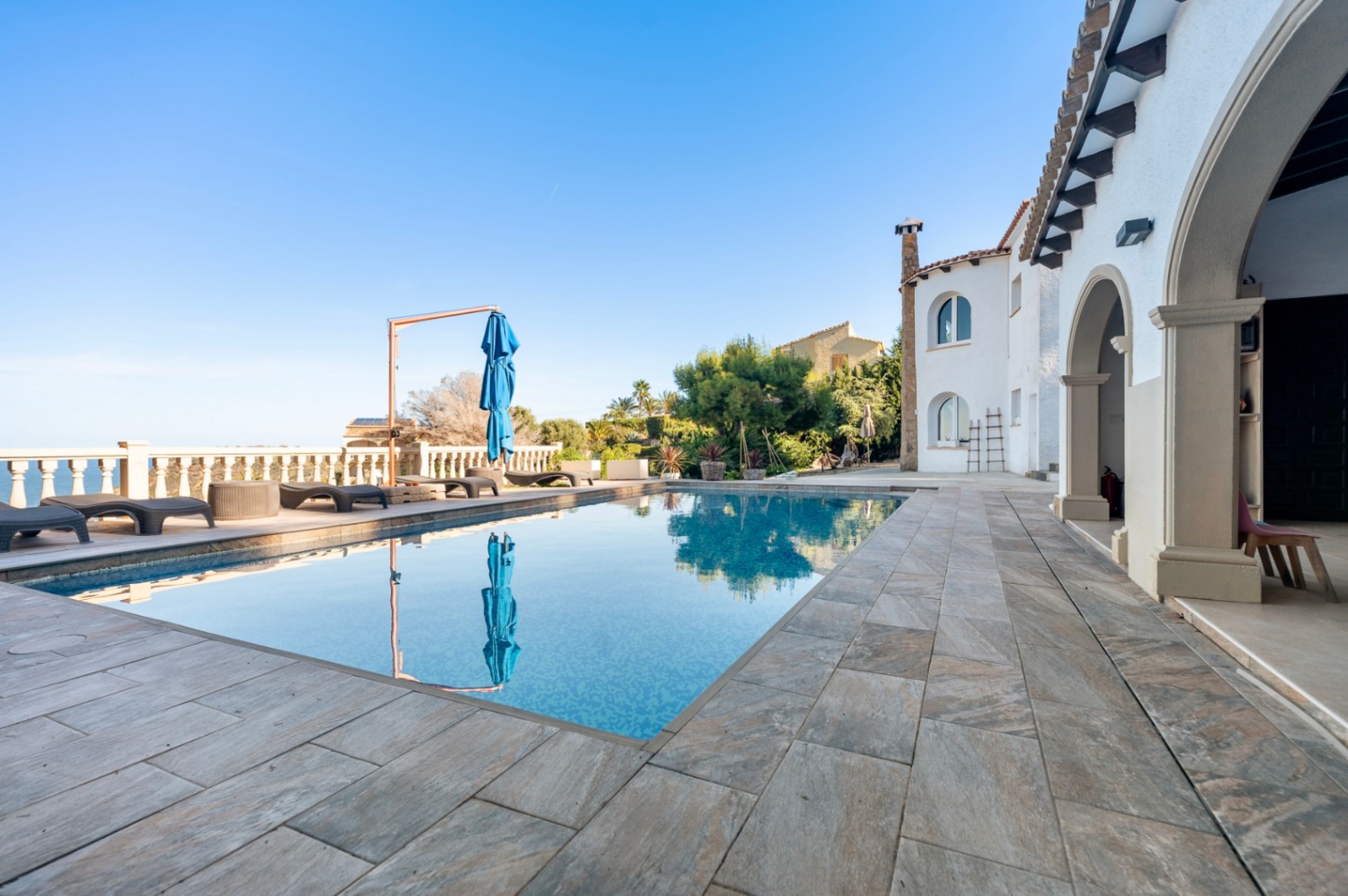 Terraza exterior con piscina rectangular, pavimentada en piedra natural y rodeada de tumbonas con vistas al mar y arquitectura tradicional de arcos.