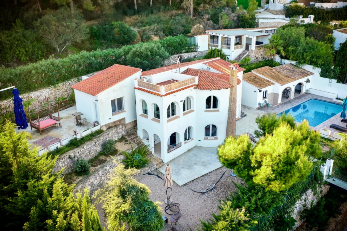 Vista aérea de la villa con fachada blanca y tejados de teja, destacando la zona de piscina, terrazas escalonadas y muros de piedra natural en un entorno boscoso.
