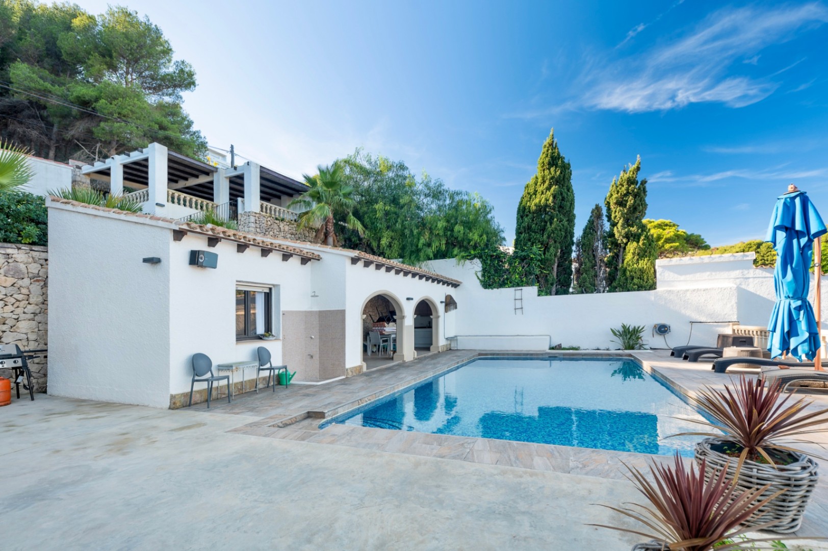 Zona de piscina rectangular con terraza de gres, ducha exterior y porche de arcos tradicionales. El entorno incluye muros de piedra, cipreses y una terraza superior con pérgola.