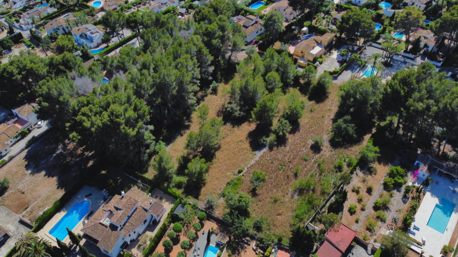 Vista aérea de la amplia parcela llana rodeada de vegetación autóctona y pinos, situada en una zona residencial consolidada con villas y piscinas privadas.