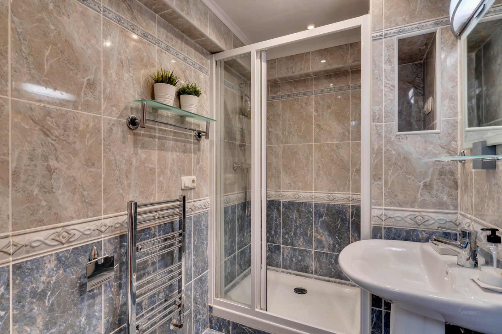 Baño con paredes alicatadas en tonos beige y azul, plato de ducha con mampara plegable, radiador toallero cromado y lavabo de pedestal blanco.