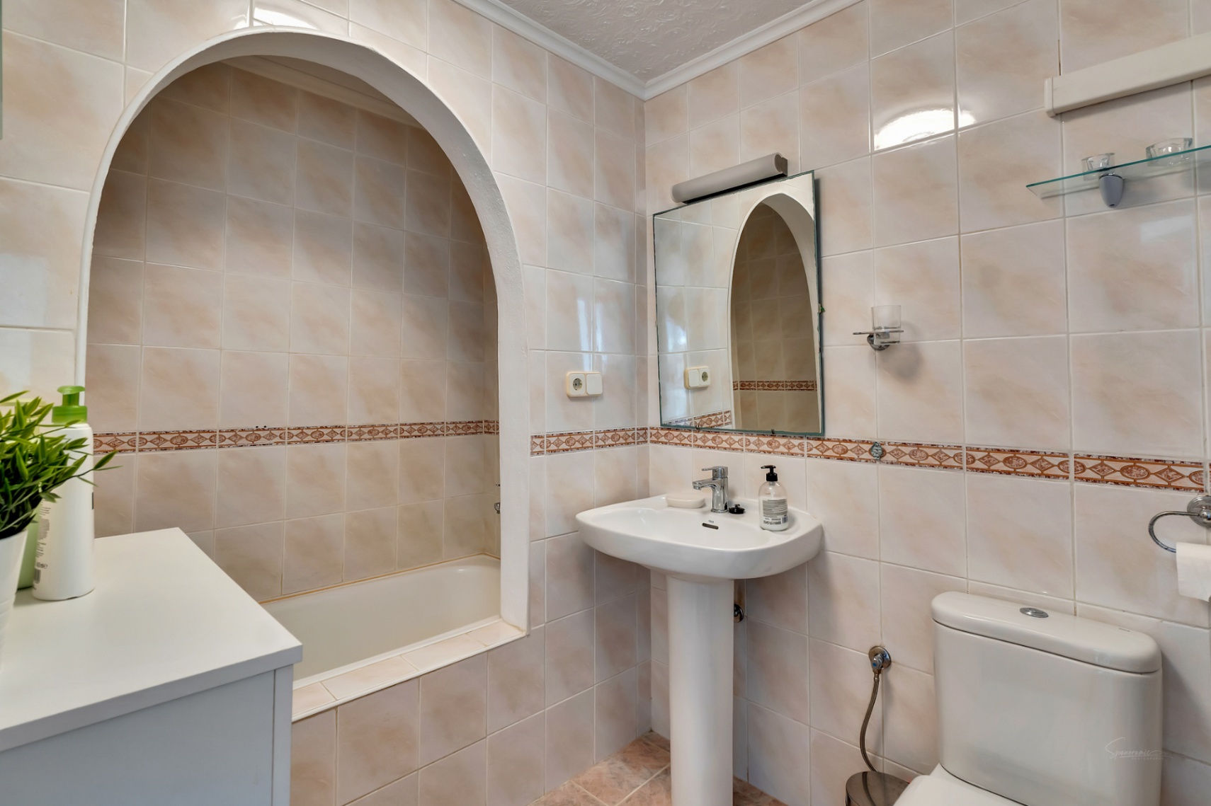 Baño de estilo clásico con bañera integrada en un arco arquitectónico, lavabo de pedestal y revestimiento cerámico en tonos crema con cenefa decorativa.