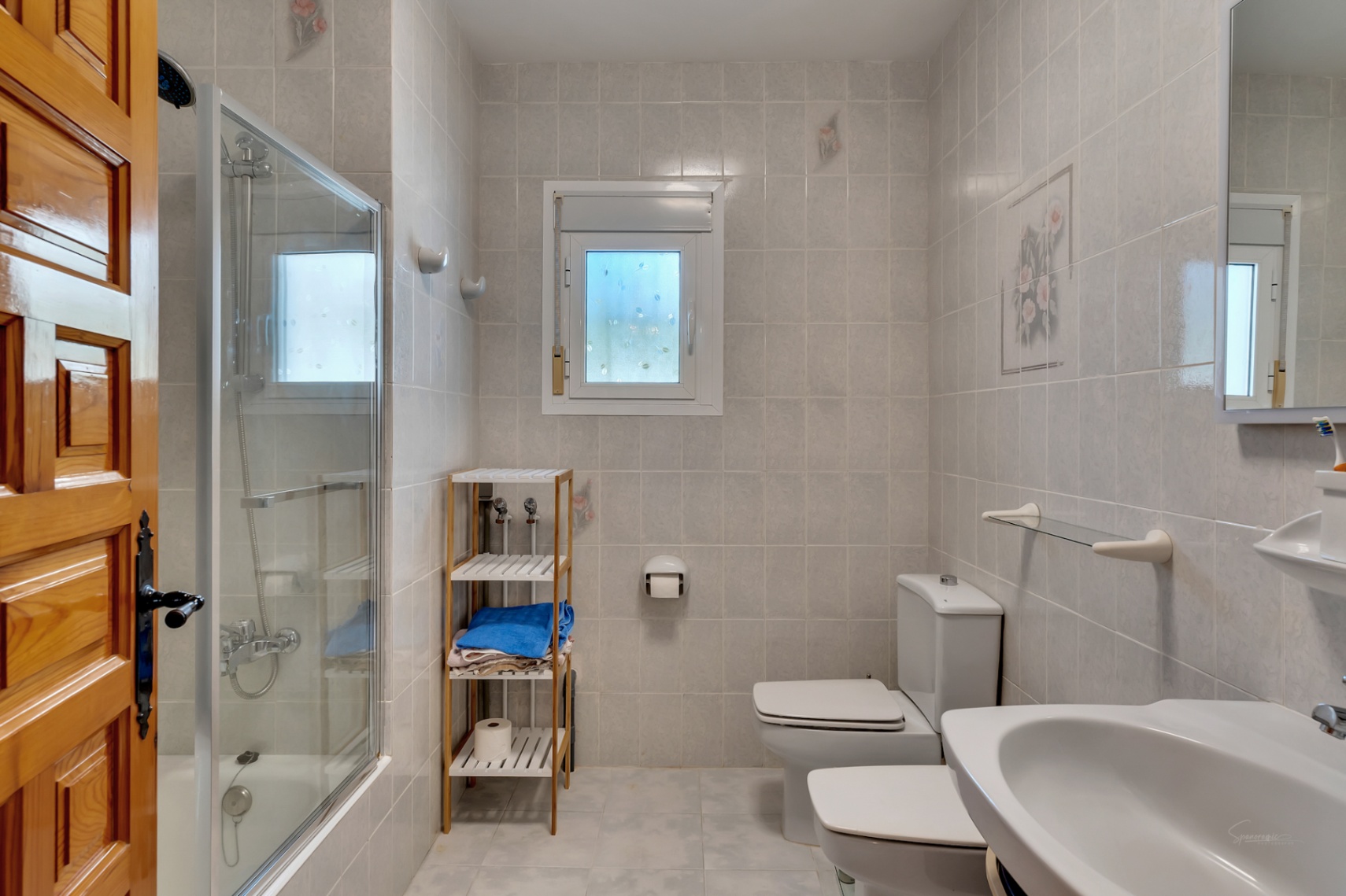 Baño funcional con ducha de mampara acristalada, sanitarios blancos y paredes alicatadas. Incluye una ventana de luz natural y puerta de madera maciza.