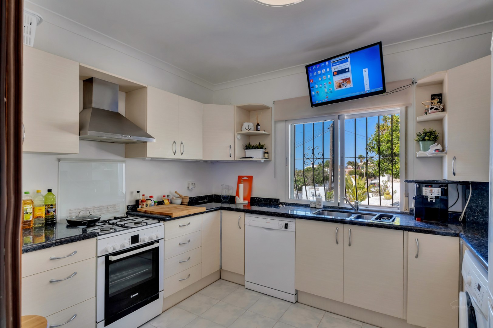 Cocina funcional equipada con encimera de granito oscuro, armarios en tono crema y electrodomésticos Bosch. Incluye ventana con rejas decorativas y televisión integrada.