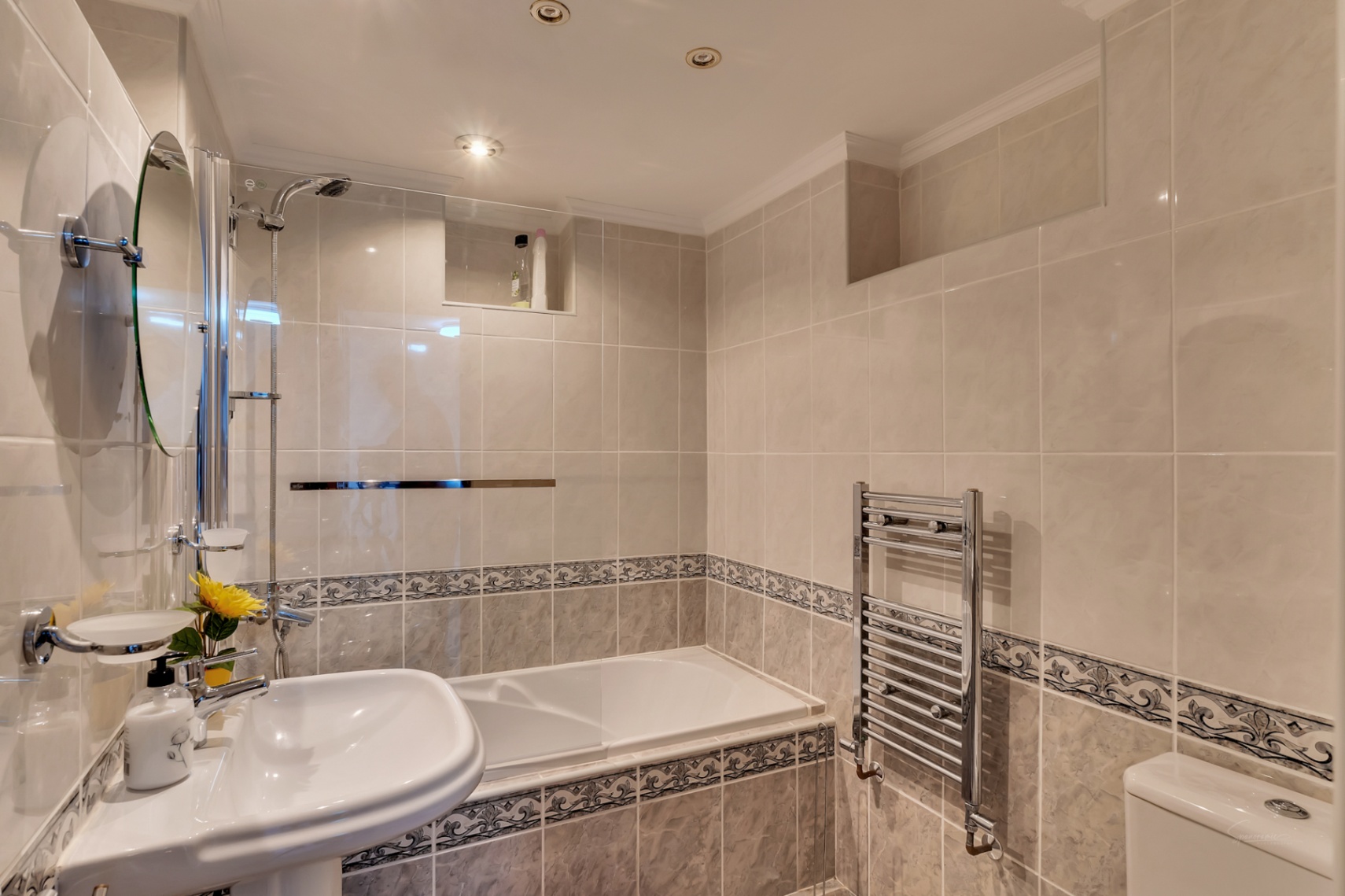 Cuarto de baño con bañera y mampara de cristal, azulejos en tonos crema con cenefa decorativa, radiador toallero cromado e iluminación empotrada en el techo.