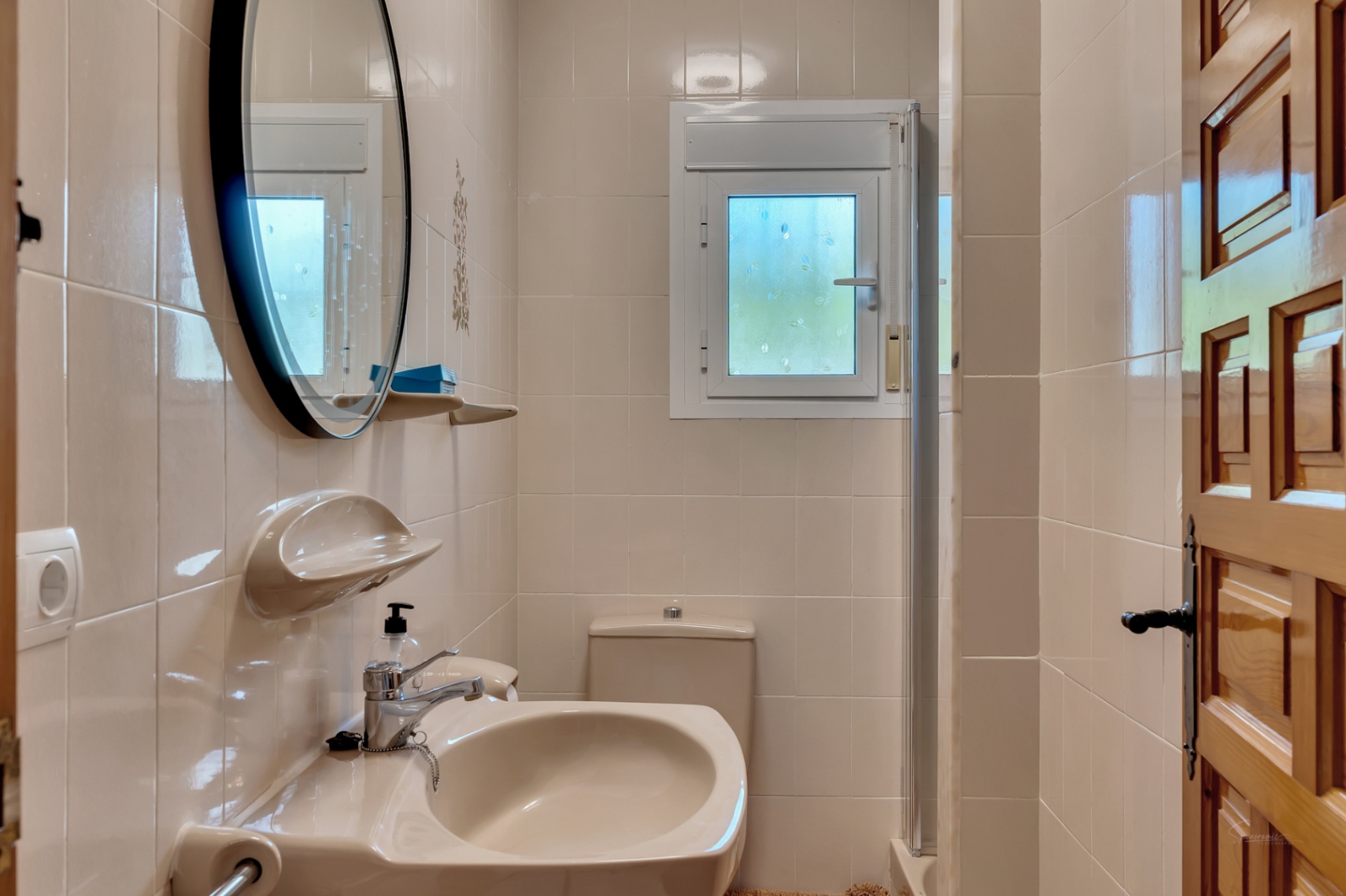 Cuarto de baño funcional con azulejos color crema, lavabo a juego, espejo circular de marco negro y ventana oscilobatiente para ventilación natural.