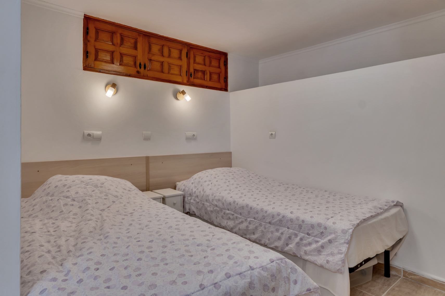 Dormitorio con dos camas individuales, cabecero de madera clara y apliques de lectura. Incluye un armario empotrado de madera de estilo tradicional en la parte superior.