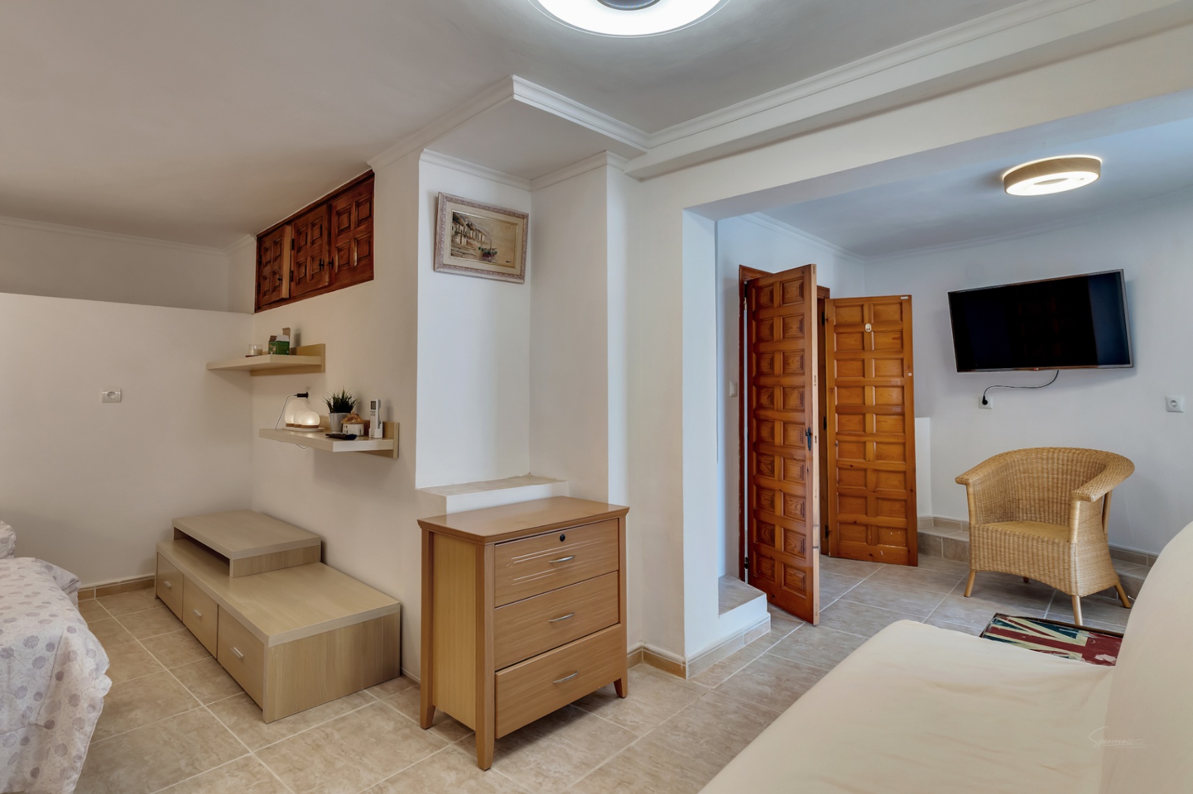 Dormitorio con suelos de gres, mobiliario de madera clara, zona de estar con sillón de mimbre y puertas rústicas de madera tallada.