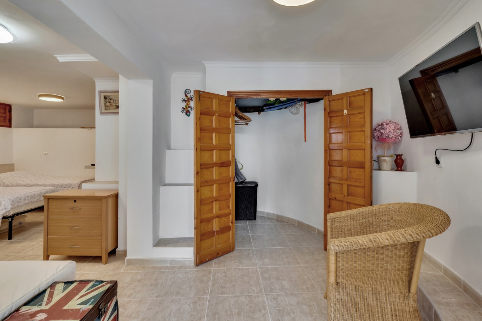 Dormitorio en planta inferior con suelos cerámicos, armario empotrado de madera y zona de estar con televisión y sillón de mimbre.