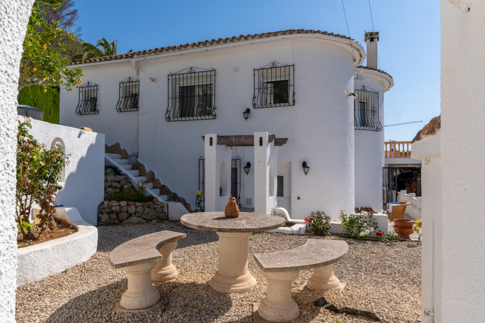 Exterior de la villa con fachada blanca y rejas de forja. El patio de grava presenta un conjunto de mesa y bancos de piedra bajo la luz natural del Mediterráneo.