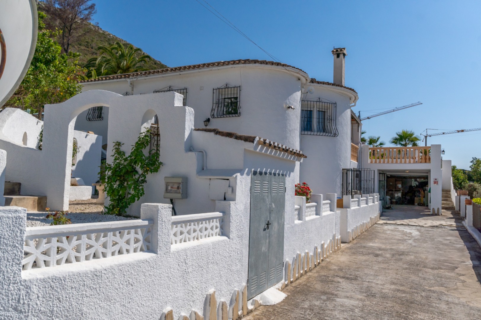 Fachada exterior de estilo mediterráneo con muros blancos, arcos arquitectónicos, rejas de forja y acceso pavimentado hacia el garaje.