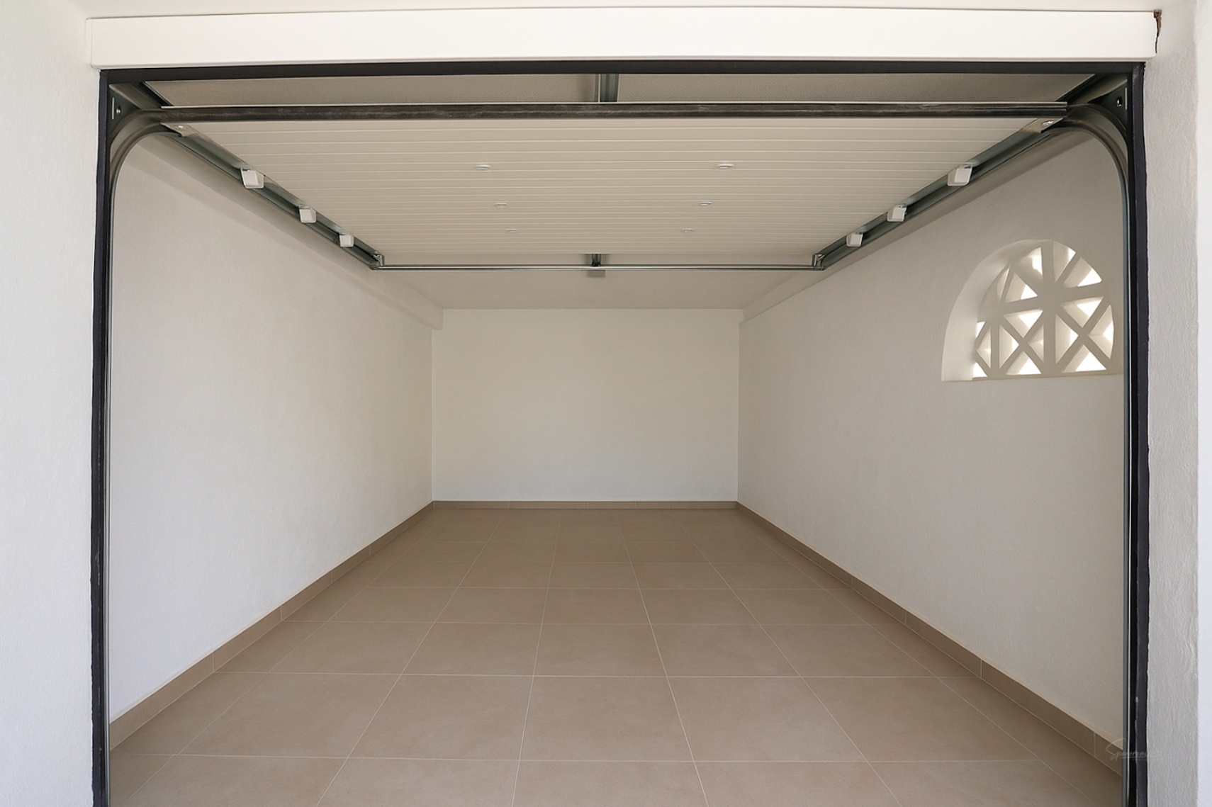Garaje privado con suelo cerámico, paredes blancas y puerta automática seccional. Incluye una ventana decorativa de arco que aporta luz natural.