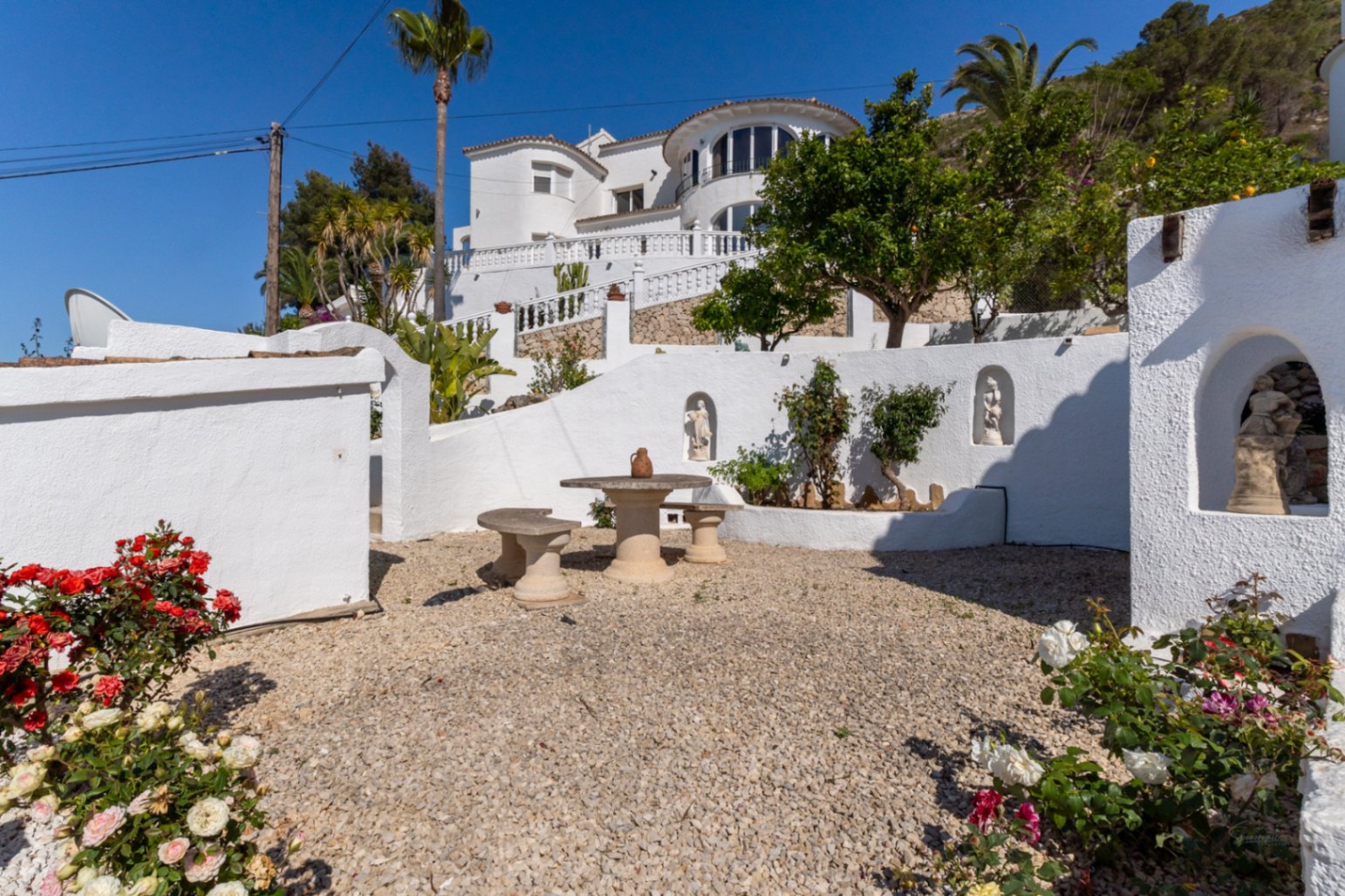 Jardín mediterráneo con suelo de grava, mesa de piedra y muros blancos decorados con hornacinas y estatuas, bajo la fachada escalonada de la villa.