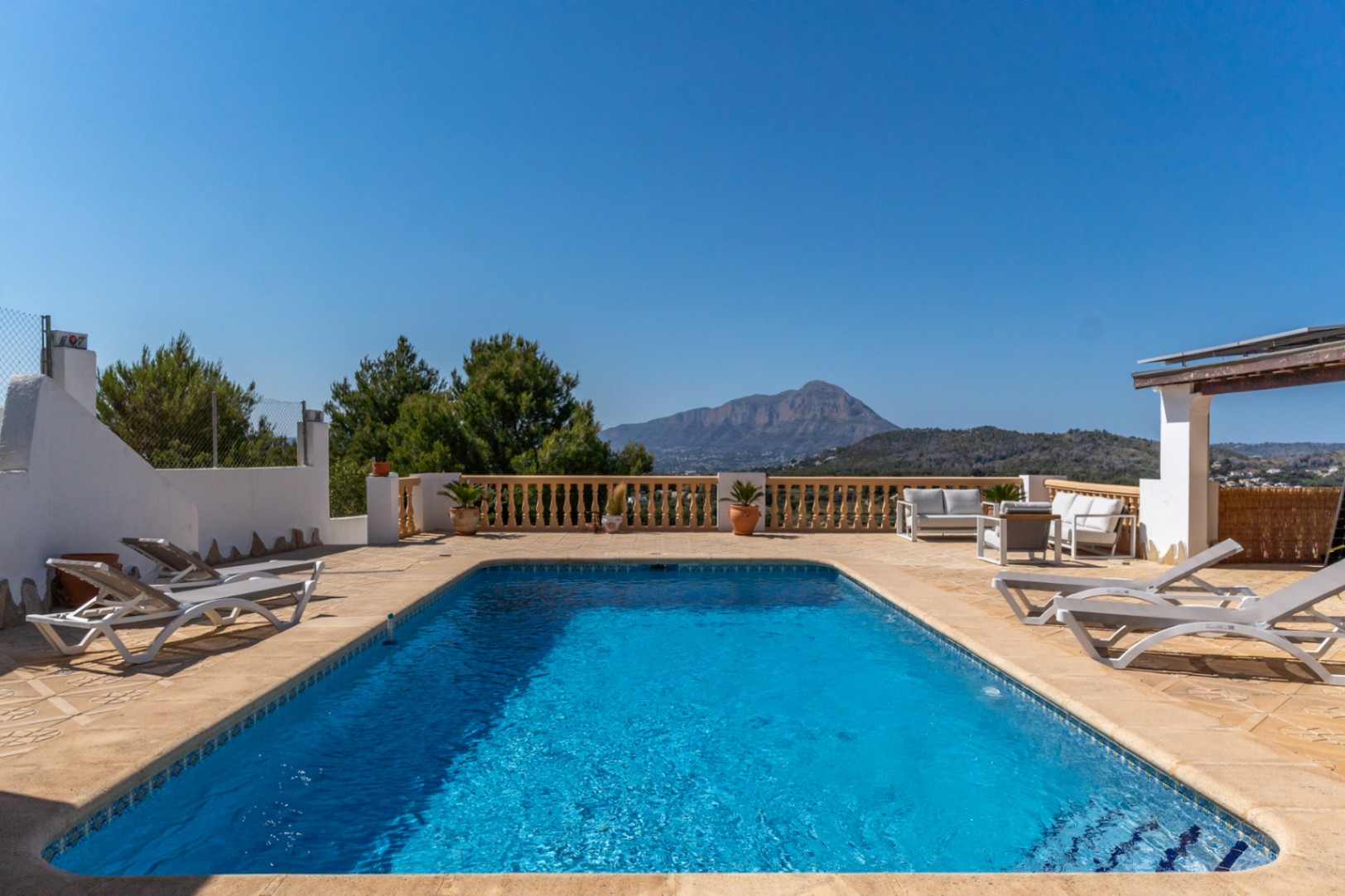 Piscina rectangular rodeada de una amplia terraza de piedra con tumbonas, zona de estar y vistas despejadas al macizo del Montgó bajo un cielo despejado.