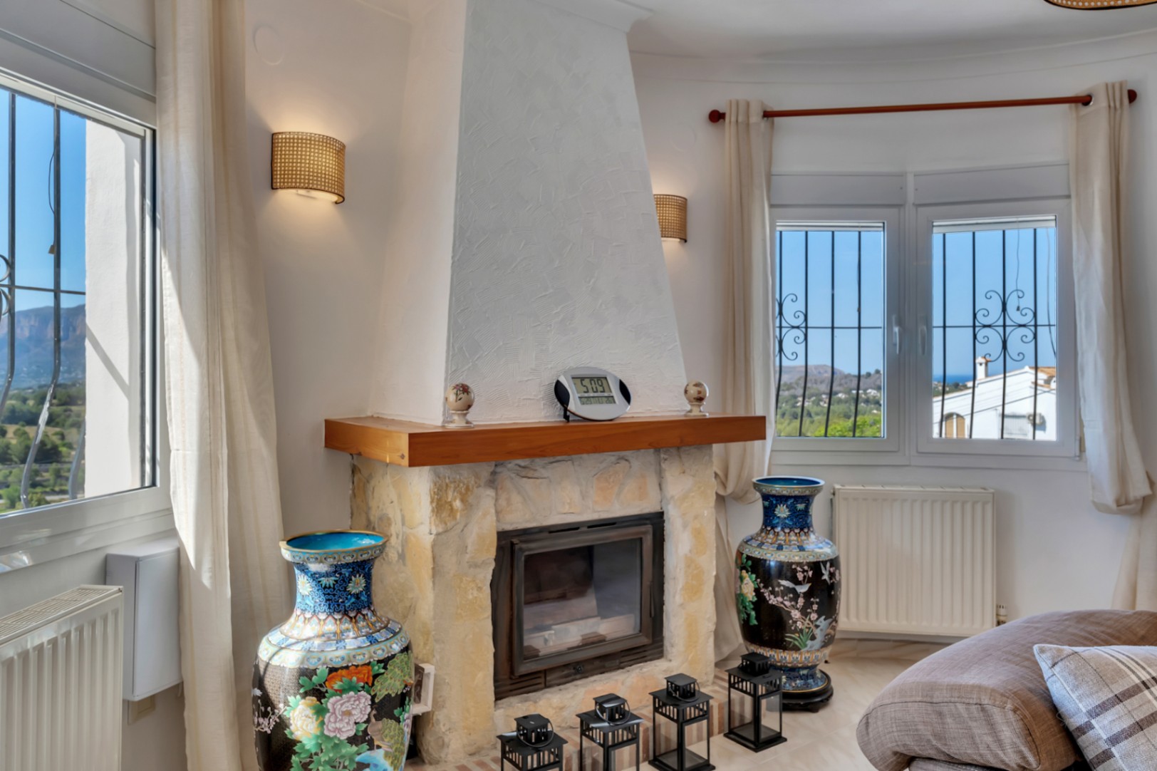 Salón con chimenea de piedra natural y repisa de madera, flanqueada por ventanales con vistas al Montgó y al mar, calefacción por radiadores y apliques decorativos.