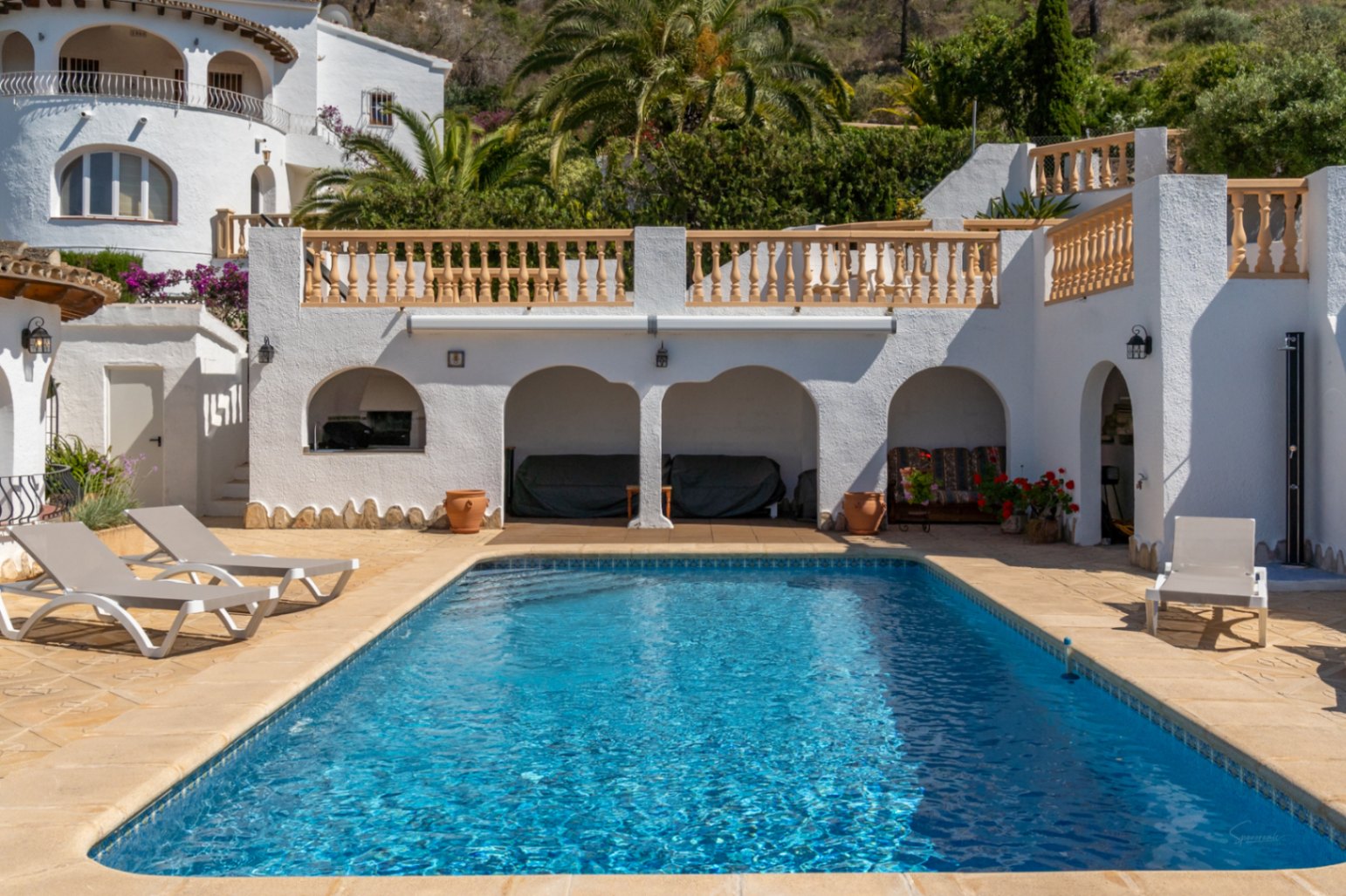 Terraza con piscina rectangular, zona de estar bajo arcos, barbacoa integrada y ducha exterior, rodeada de arquitectura blanca de estilo mediterráneo.