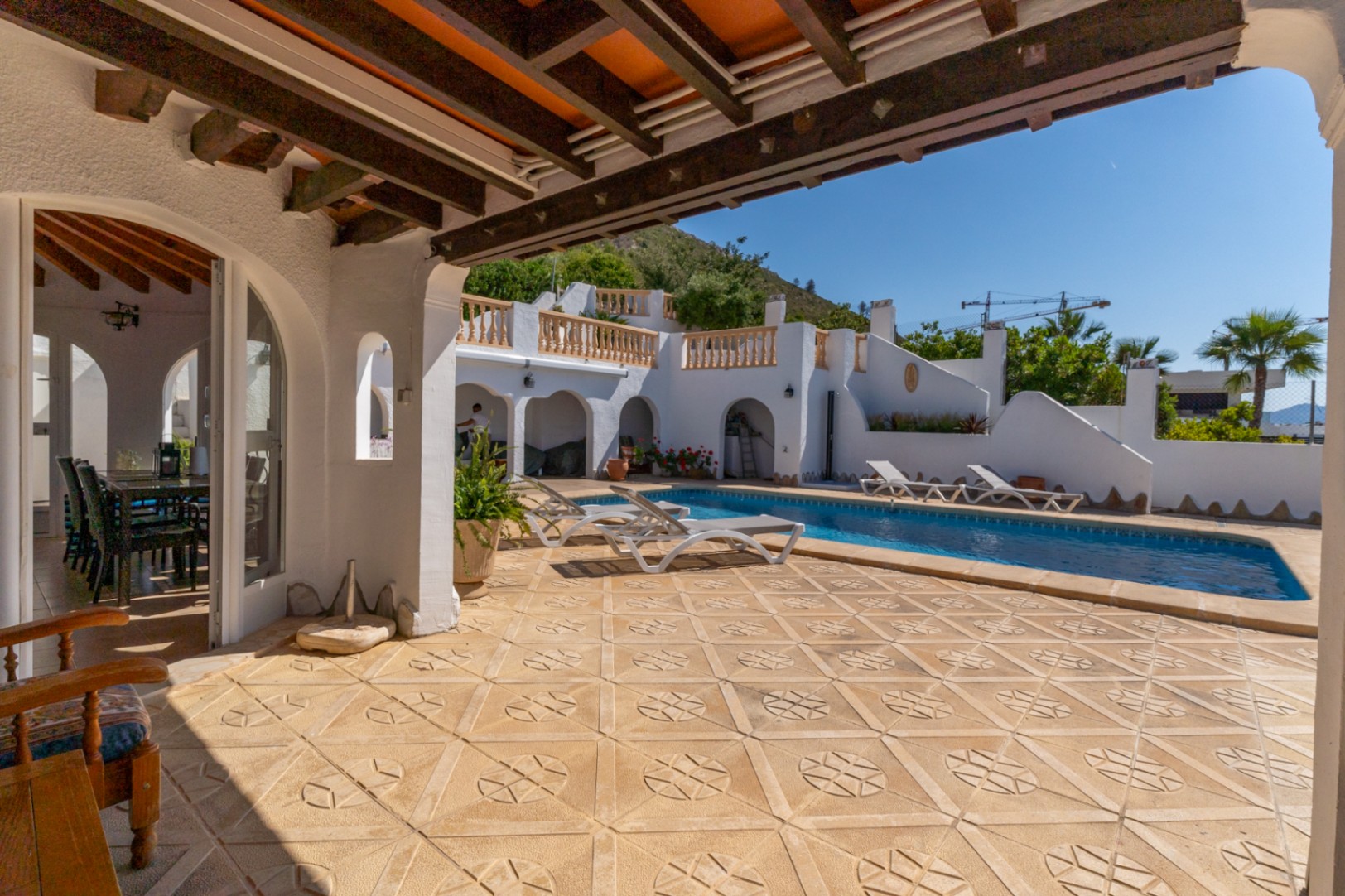 Terraza exterior con suelos de piedra grabada, piscina rectangular y porche cubierto con vigas de madera vista y arcos de estilo mediterráneo.