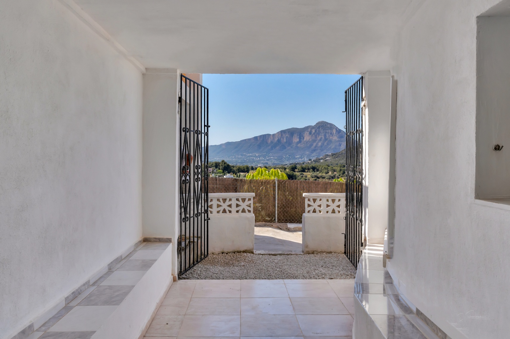 Vista panorámica del Montgó desde el porche de entrada con paredes blancas, suelos cerámicos y puertas de hierro forjado que enmarcan el paisaje natural.