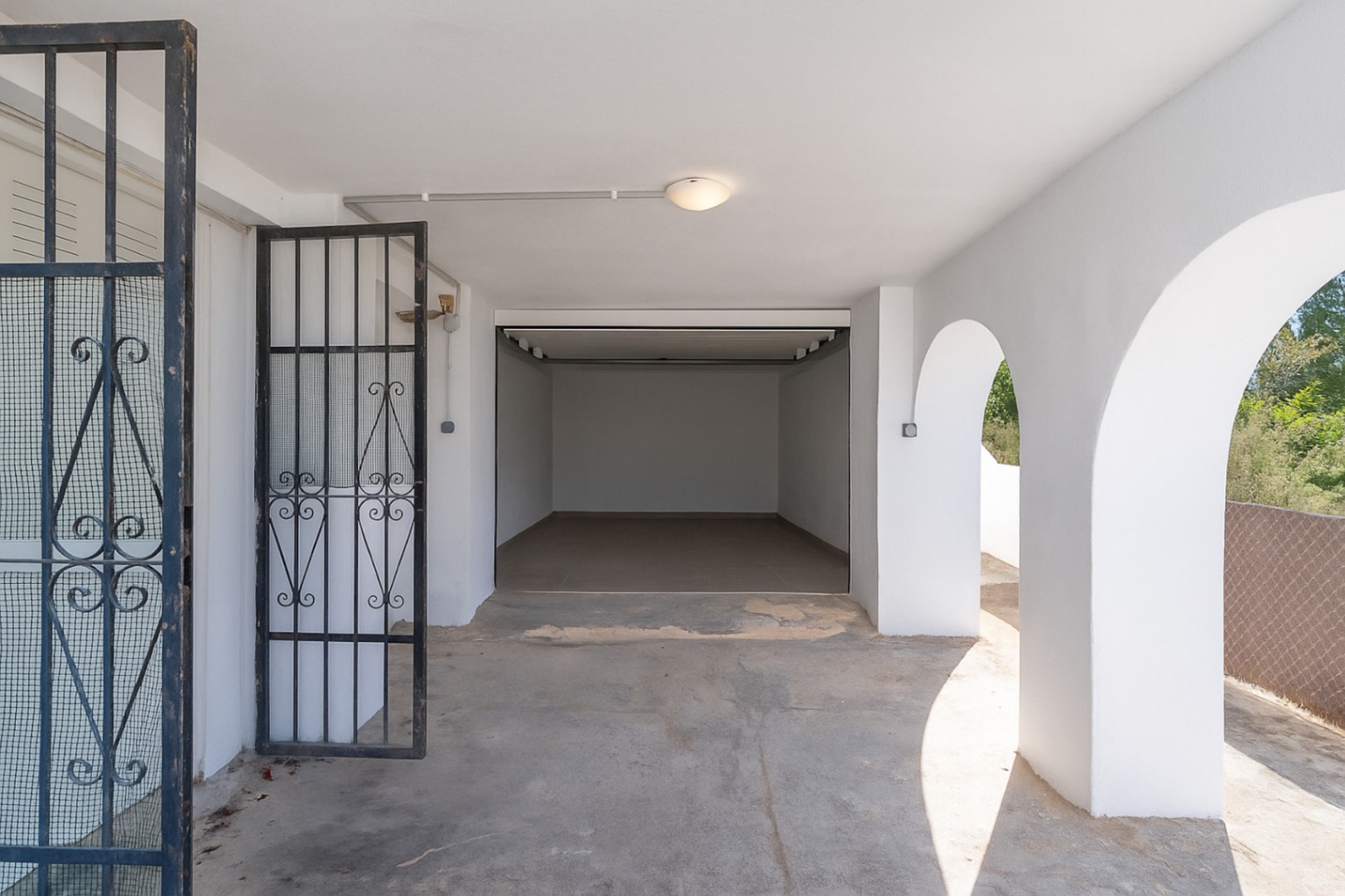 Zona de aparcamiento cubierta con arcos mediterráneos blancos, acceso mediante puerta de forja y garaje cerrado con suelo cerámico.