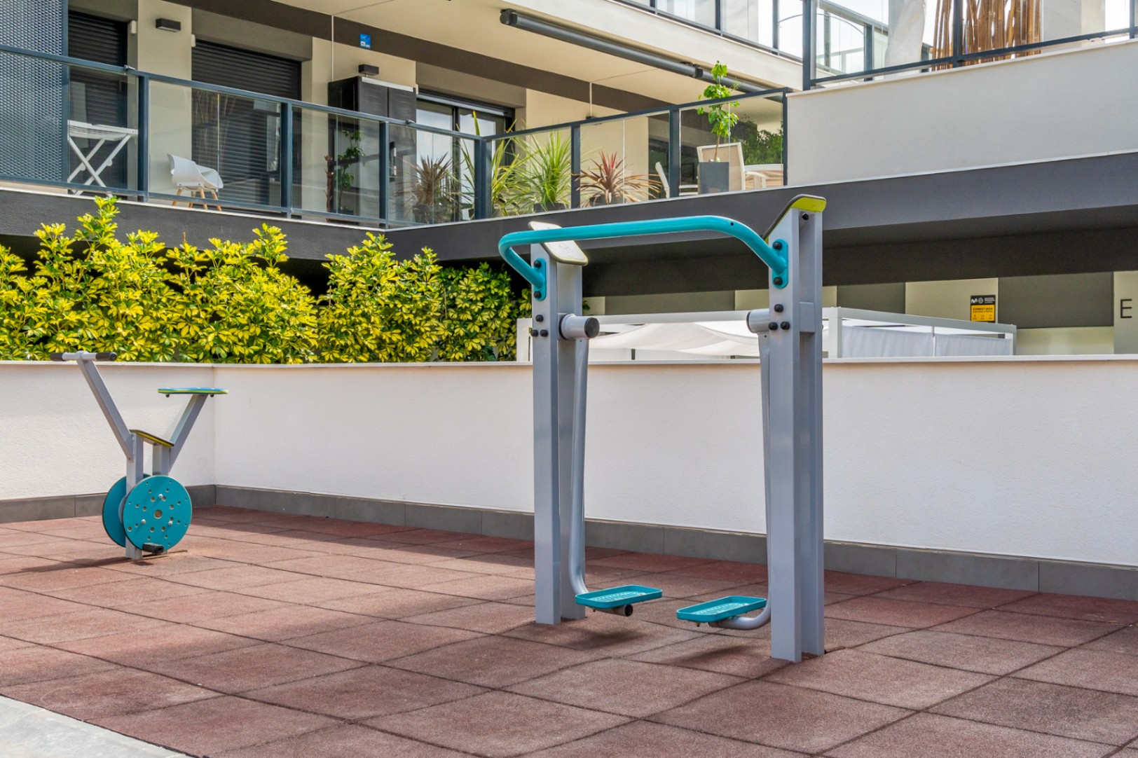 Área de gimnasio exterior equipada con máquinas de cardio sobre suelo técnico de seguridad, integrada en las zonas comunes del complejo moderno.