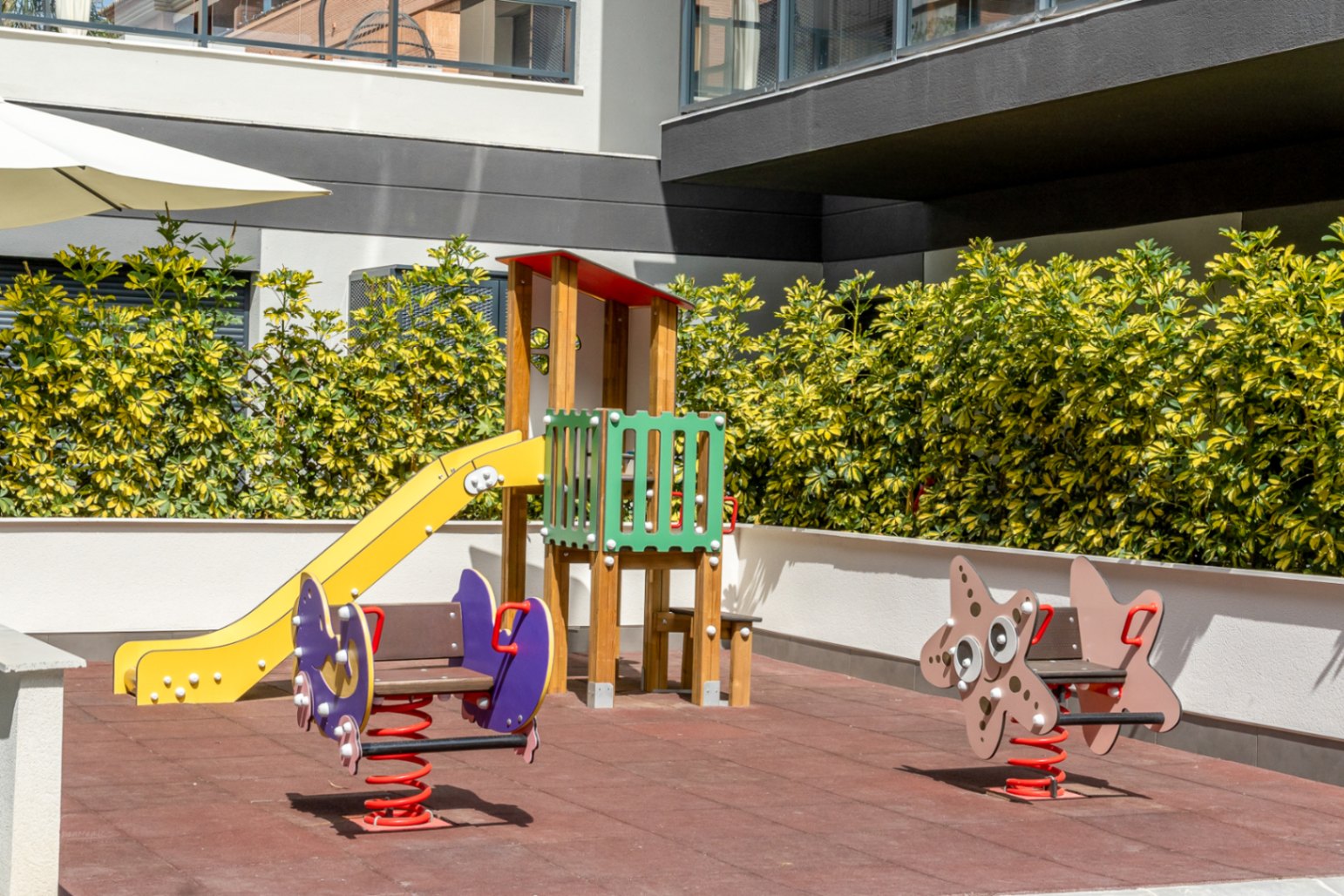 Área de juegos infantiles con suelo de seguridad, tobogán de madera y balancines de muelle, rodeada de setos cuidados en la zona comunitaria.