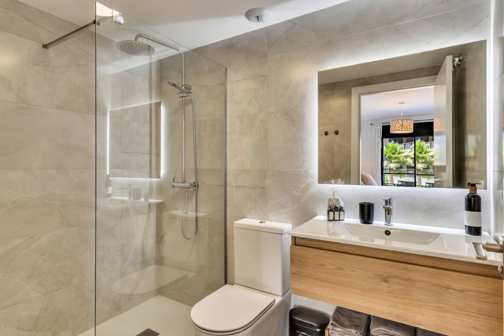 Baño moderno con ducha italiana, mampara de cristal y grifería cromada. Destaca el mueble de lavabo suspendido en madera y el espejo con retroiluminación LED perimetral.
