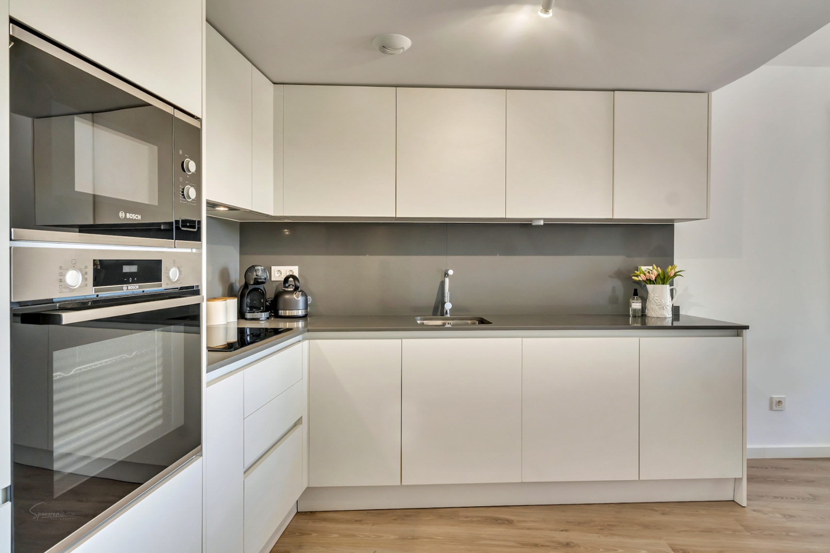 Cocina moderna de diseño minimalista con armarios blancos sin tiradores, encimera gris y electrodomésticos Bosch integrados sobre suelo de madera clara.